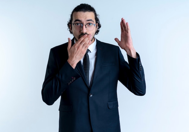 Business man in black suit and glasses raising hand looking surprised covering mouth with hand standing over white wall