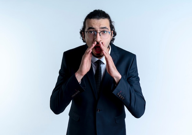 Business man in black suit and glasses shouting or calling with hands near mouth standing over white wall