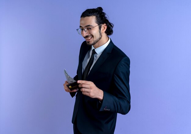 Business man in black suit and glasses showing cash and credit card smiling cheerfully standing over blue wall