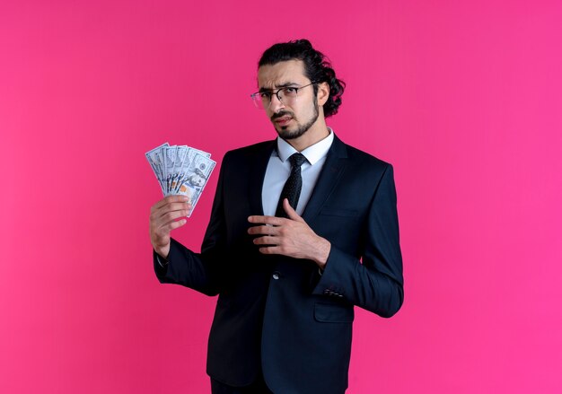 Business man in black suit and glasses showing cash looking to the front confused and displeased standing over pink wall