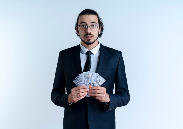 Business man in black suit and glasses showing cash looking to the front confused and very anxious standing over white wall