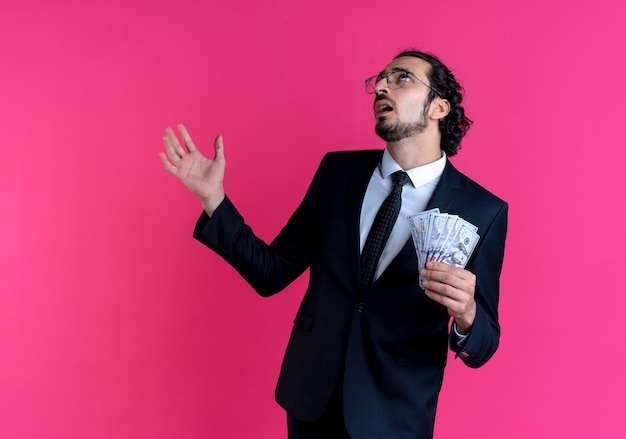 Business man in black suit and glasses showing cash, looking up confused with arm out standing over pink wall