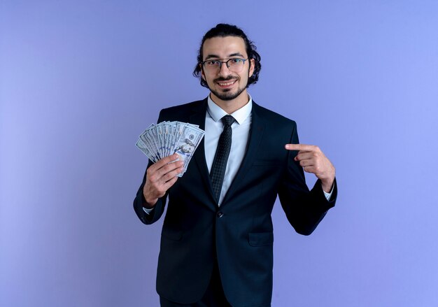 Деловой человек в черном костюме и очках показывает деньги, указывая пальцем на него, весело улыбаясь, стоя над синей стеной