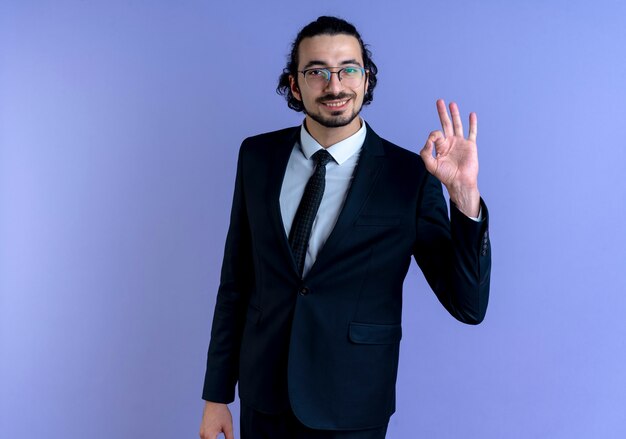 검은 양복과 파란색 벽 위에 유쾌하게 서있는 정면을보고 확인 기호를 보여주는 안경 비즈니스 남자