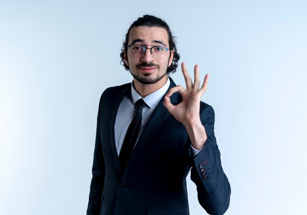 Business man in black suit and glasses showing ok sign smiling confident standing over white wall