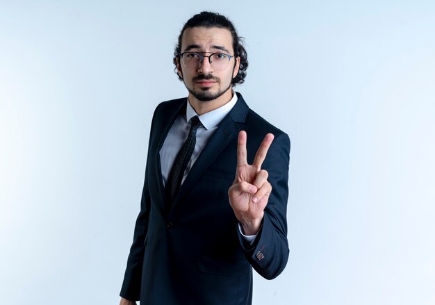 Business man in black suit and glasses showing and pointing up with fingers number two looking with serious face standing over white wall
