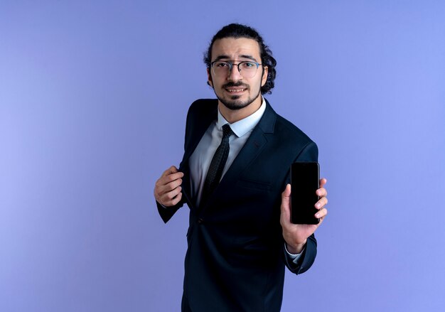 Business man in black suit and glasses showing smartphone looking to the front confused standing over blue wall