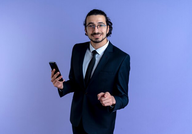 Business man in black suit and glasses showing smartphone pointing with finger to the front smiling confident standing over blue wall