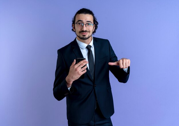 Business man in black suit and glasses showing smartphone pointing with finger to it smiling confident standing over blue wall