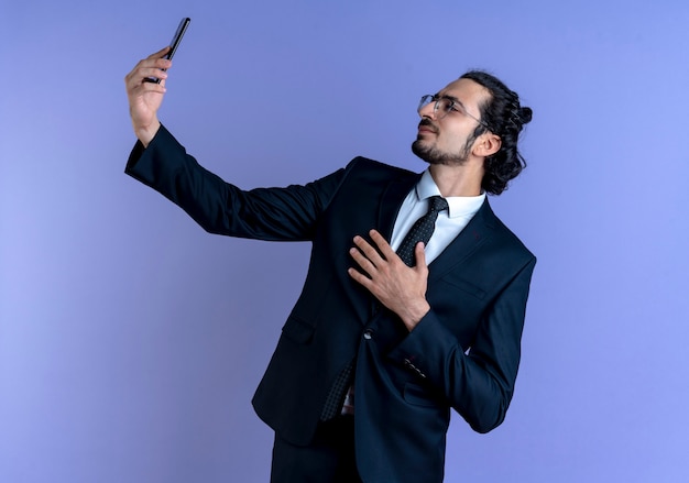 검은 양복과 파란색 벽 위에 자신감이 서있는 자신의 스마트 폰을 사용하여 셀카를 복용하는 안경 비즈니스 남자