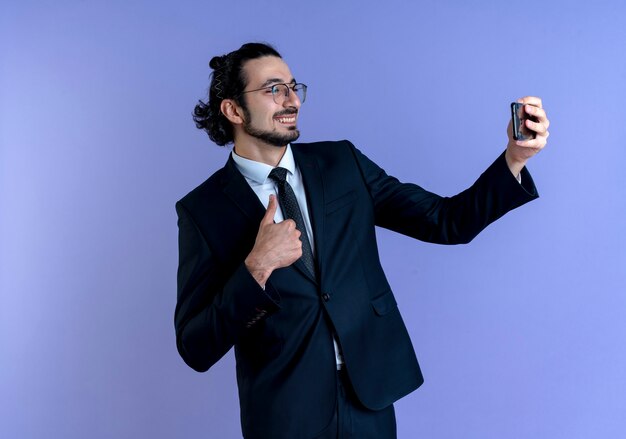 검은 양복과 파란색 벽 위에 서 엄지 손가락을 보여주는 스마트 폰을 사용하여 셀카를 복용하는 안경 비즈니스 남자