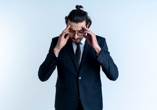 Business man in black suit and glasses touching his temples suffering from headache standing over white wall