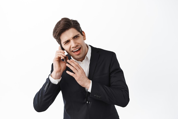 Business man cant hear person on the phone having a call crowded loud place frowning confused standing in black suit against white background Copy space