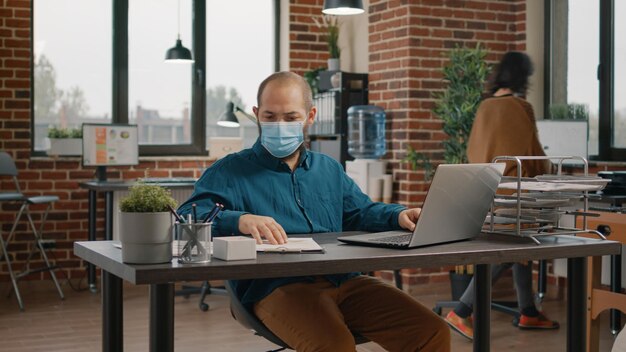 Business man doing paperwork and looking at rate charts while working with laptop. Employee wearing face mask and analyzing data analysis on files for project planning and strategy.