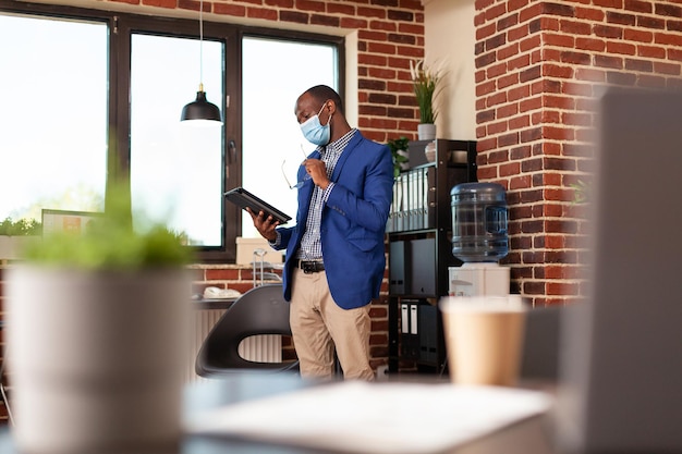 Business man wearing face mask and using digital tablet to plan company project. Employee standing in startup office and working on device with technology during coronavirus pandemic.