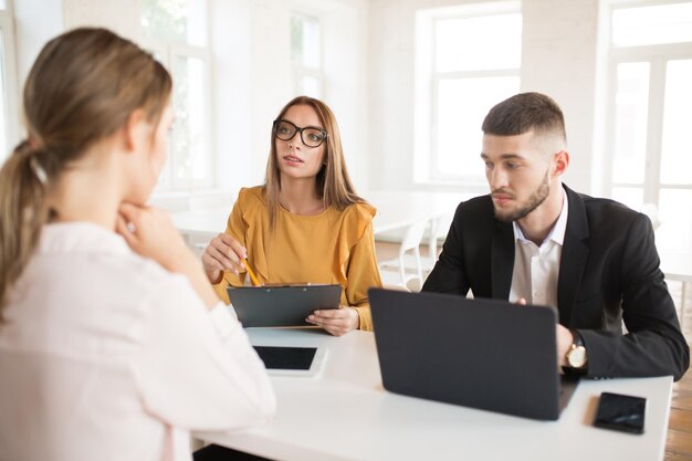 Business man with laptop thoughtfully looking aside while business woman in eyeglasses with folder in hand talking with applicant about work Young employers spending job interview in modern office