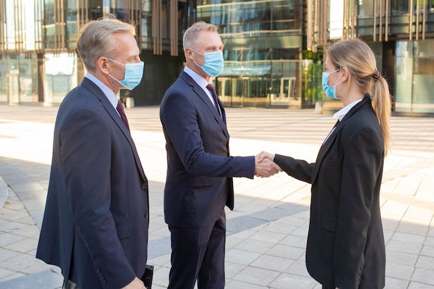 Business men and woman in face masks and office suits meeting in city, shaking hands near building. Side view shot. Communication and virus protection concept