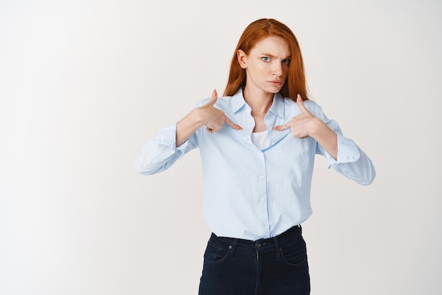 Business people Confused redhead female pointing at herself and looking with disbelief at camera standing puzzled against white background