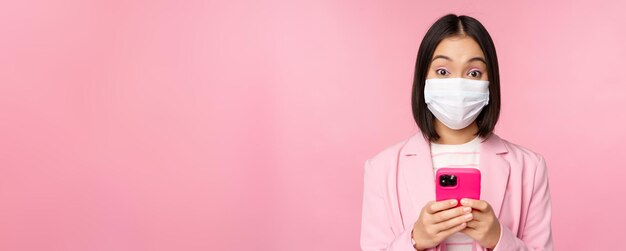 Business people and covid19 concept Young asian businesswoman in suit and medical face mask using smartphone app standing over pink background