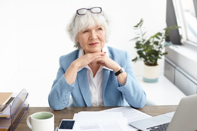 Business, people, electronic gadgets and modern technology concept. Indoor shot of experienced senior Caucasian female CEO in formal wear having pensive look while working at desk with laptop