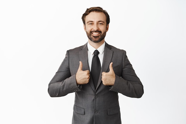 ビジネスマン笑顔で親指を立てて賞賛を示すスーツを着たハンサムな企業の男性は、白い背景の上に立ってはいと言って良い仕事をしています