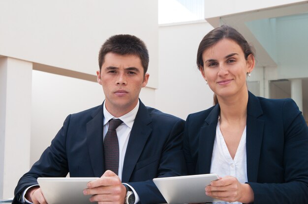 Business people looking at camera and holding tablets at desk