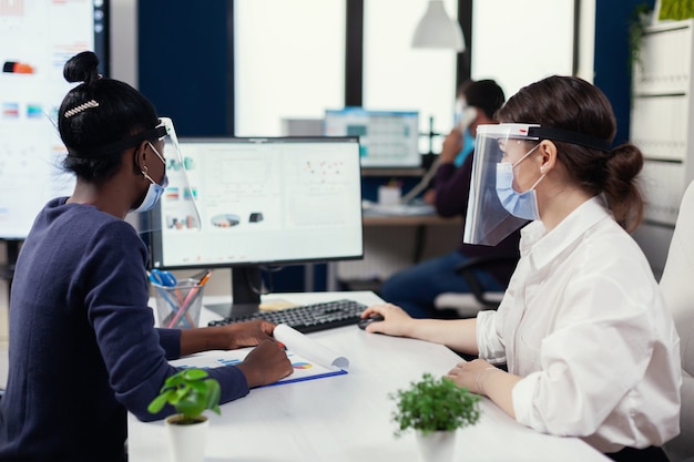 Business people using computer with financial graph wearing face mask for covid19. Multiethnic team working in company with new normal respecting social distance during global pandemic with coronaviru