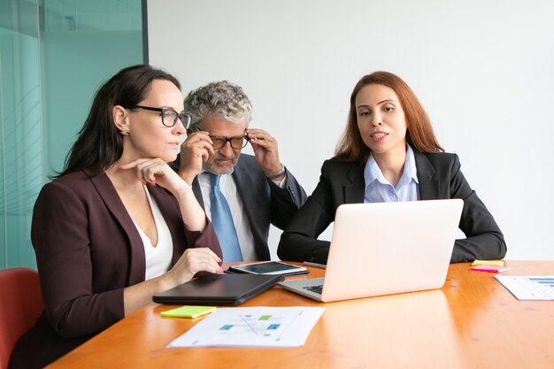 Business people watching project presentation on laptop, sitting at meeting table with paper reports and charts.