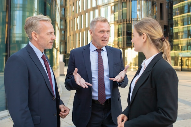 Free photo business team discussing project outdoors. men and woman wearing office suits, standing and talking outdoors with city building in background. meeting and cooperation concept