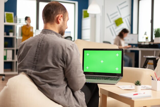 Free photo business team sitting on couch analysing financial statistics, holding laptop with green screen while diverse team working on background