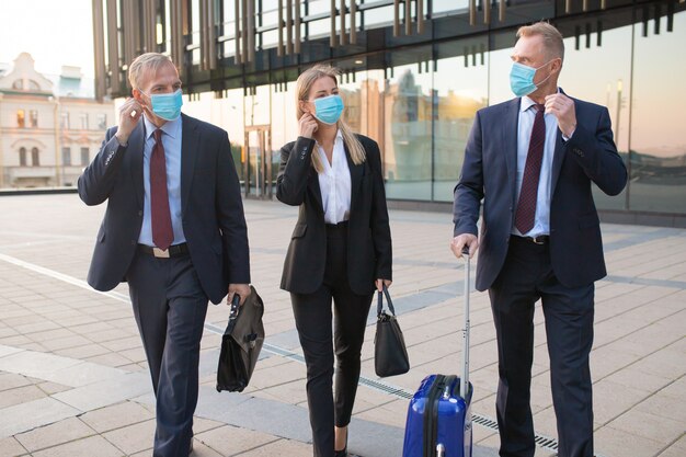Business tourists in face masks travelling with briefcases or suitcase, walking outdoors, talking to each other. Front view. Business trip and epidemic concept