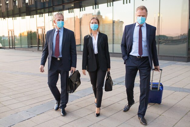 Business tourists in face masks visiting foreign partner office, wheeling suitcase, walking outdoors. Front view. Business trip and epidemic concept