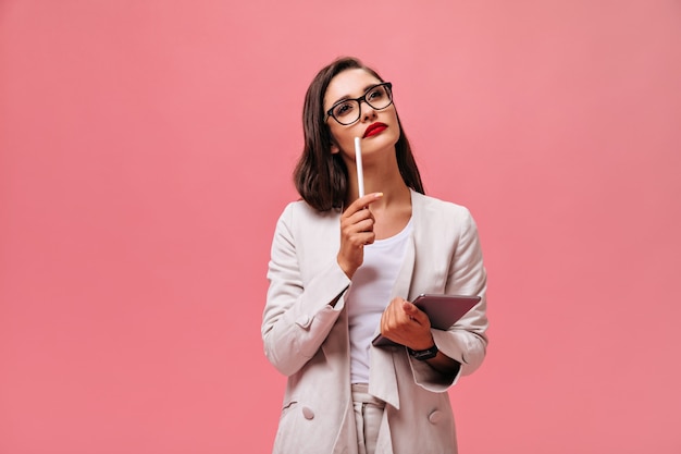 Business woman in beige suit thoughtfully posing on pink background.  Pensive girl in light stylish outfit holds tablet on isolated backdrop.