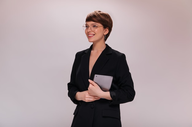 Business woman in black suit holds computer tablet. Short-haired employee in dark jacket smiles widely on isolated backdrop