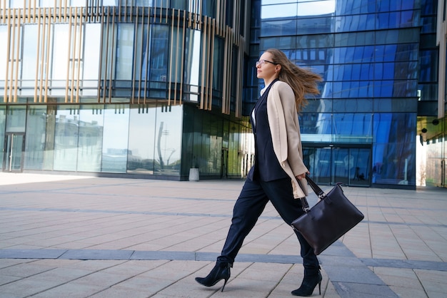 A business woman in a coat and suit, holding a bag in her hand, walks near the business center during the day.