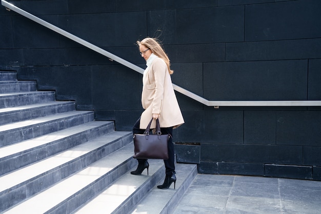 그녀의 손에 가방과 코트를 입은 비즈니스 우먼은 건물의 계단을 올라갑니다.