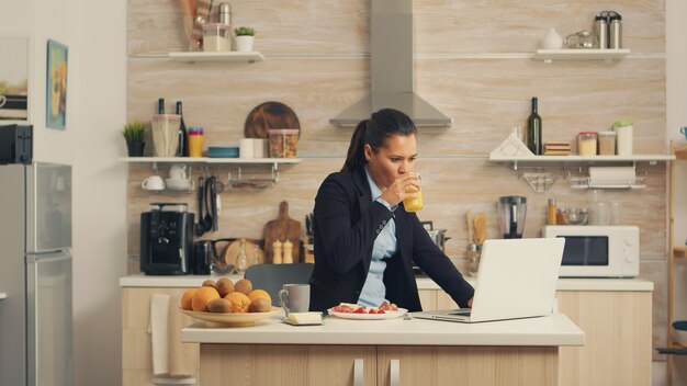 Business woman eating breakfast and working on laptop. Concentrated business woman in the morning multitasking in the kitchen before going to the office, stressful way of life, career and goals to mee