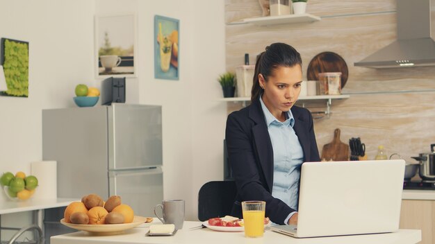 아침 식사 중에 노트북 작업을 하는 동안 버터와 함께 구운 빵을 먹는 비즈니스 여성. 사무실에 가기 전에 부엌에서 멀티태스킹을 하는 아침에 집중된 비즈니스 우먼, 스트레스 w