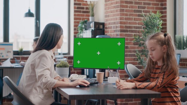 Business woman looking at green screen while daughter drawing on paper at workplace desk. Working mother using isolated template and mockup background with chroma key, having child at work