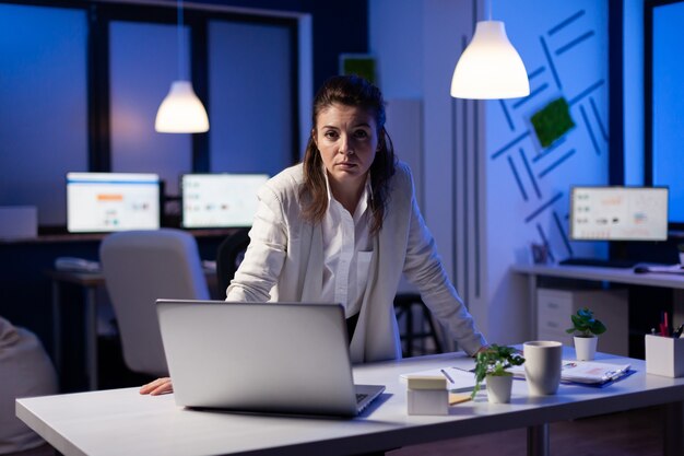 Business woman looking tired at camera standing near desk in start-up business company late at night