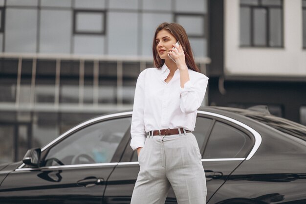 ビジネスの女性が車のそばに立っていると電話を使用して