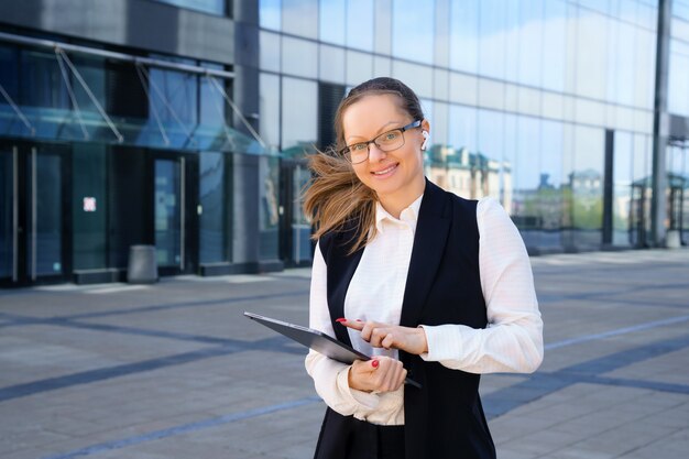 A business woman stands with a laptop in a suit and glasses outside an office building during the day.