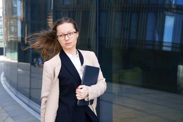 Деловая женщина стоит с ноутбуком в костюме и очках возле офисного здания днем.