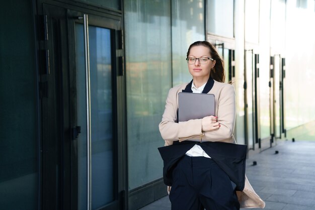 A business woman stands with a laptop in a suit and glasses outside an office building during the day.