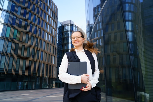 A business woman stands with a laptop in a suit and glasses outside an office building during the day.