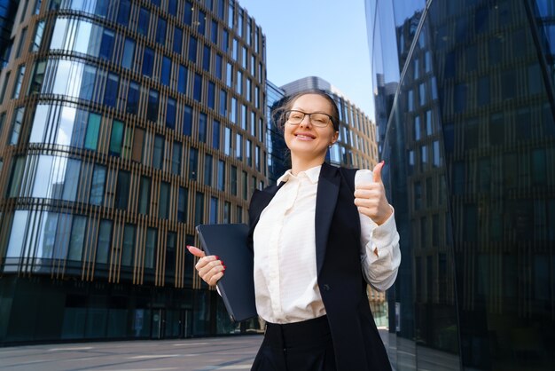 A business woman stands with a laptop in a suit and glasses outside an office building during the day.