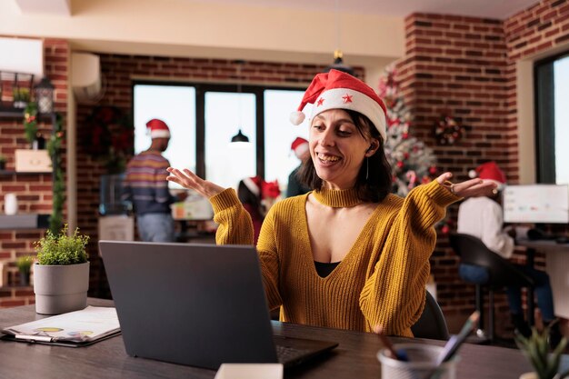 Business woman talking on videocall conference in office with christmas tree and festive decorations. Attending remote teleconference call and online meeting during holiday season at work.