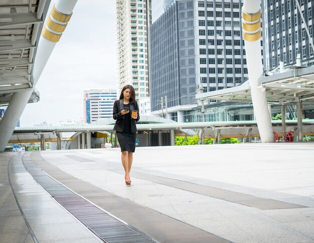 Business woman using phone with coffee in hand walking on the street with office buildings in the background