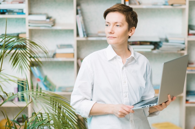 business woman with laptop in a spacious study with a palm tree and shelves looking 