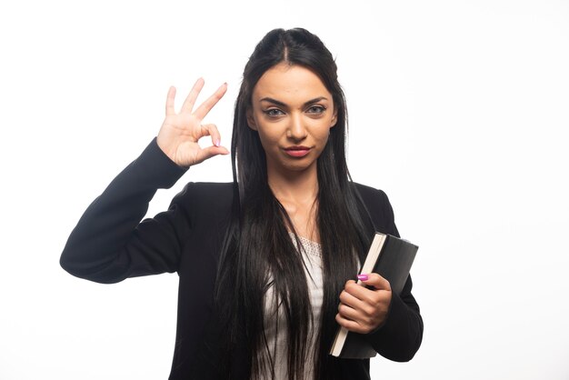 Business woman with ok gesture holding clipboard on white wall